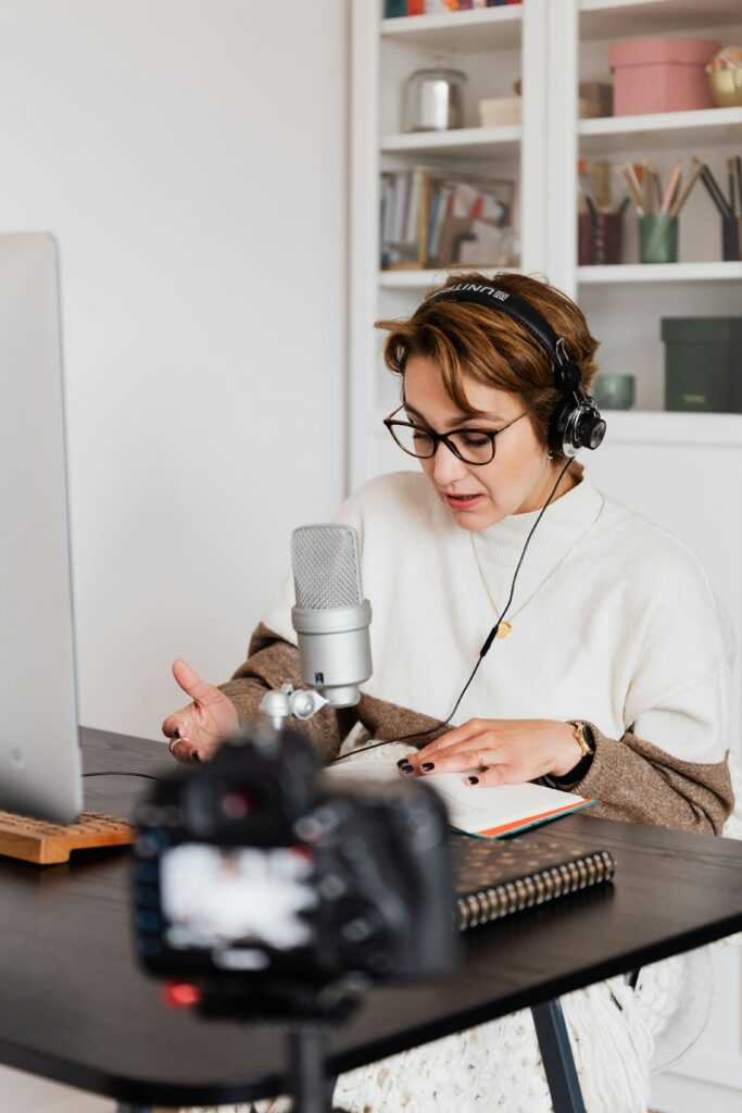 Woman wearing headphones narrating an audiobook at a desk with a microphone, computer, and notes.